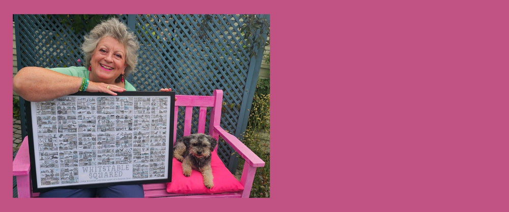Caroline Wagstaff the Artist holding a print of Whitstable Squared in a black frame sitting on a pink bench with her dog Boots.